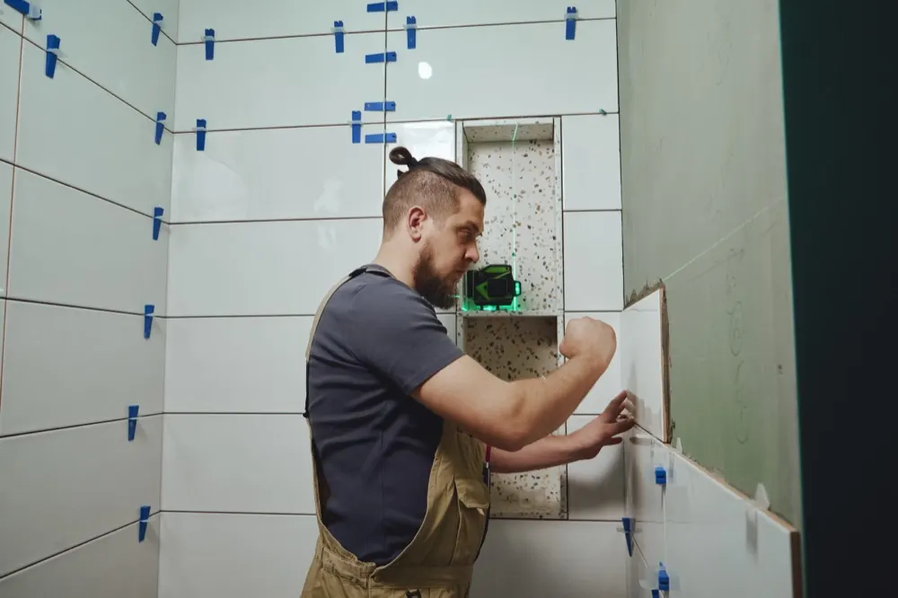 A tiler setting bathroom wall tiles with levelling clips and a laser level.