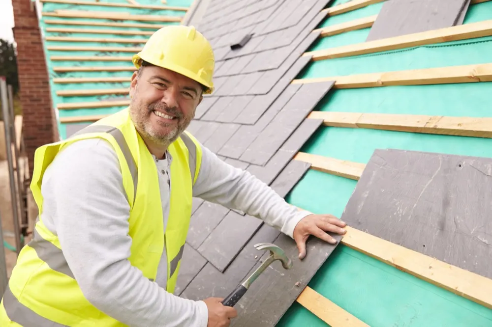 A roofer in a hard hat and hi-vis laying slates on a battened roof.