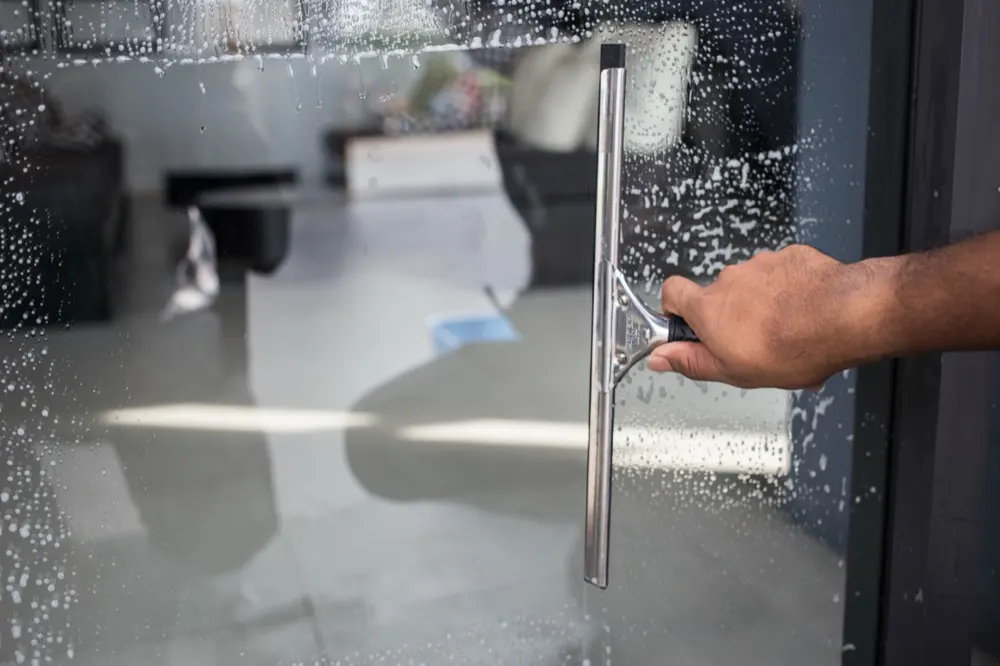A squeegee being drawn down a soapy glass door, a room visible through it.